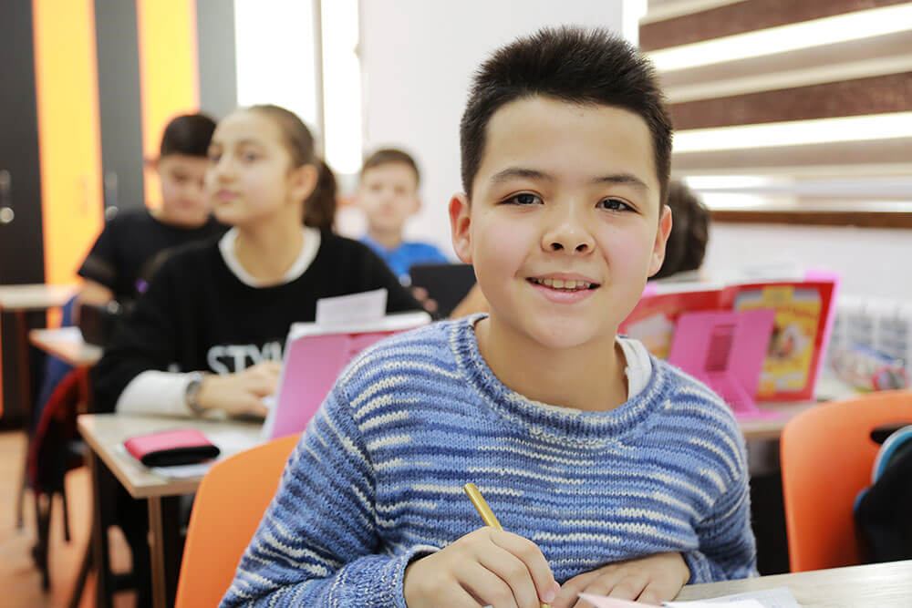 Boy smiling at camera while sitting in active classroom alongside other students